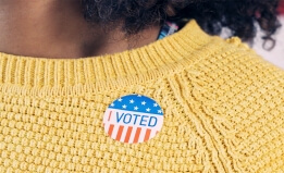 Cropped photograph of a person wearing a yellow sweater with an 'I Voted' sticker